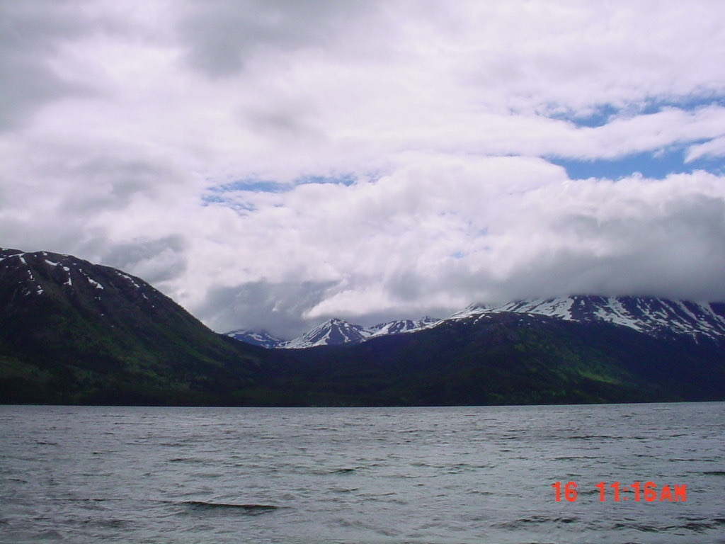 Auto-generated description: Snow-capped mountains are partially covered by clouds above a large body of water under a partly cloudy sky.