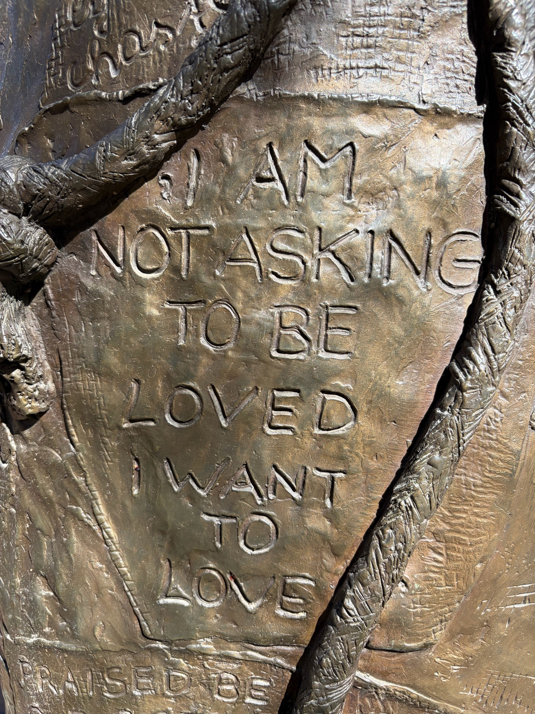 A close-up of a bronze sculpture with text carved into its surface reading: “I am not asking to be loved. I want to love.” Partial text is also visible at the top (“chas…”) and bottom (“Praised be”). The surface has an organic, textured quality with intertwining forms surrounding the inscribed panel.​​​​​​​​​​​​​​​​