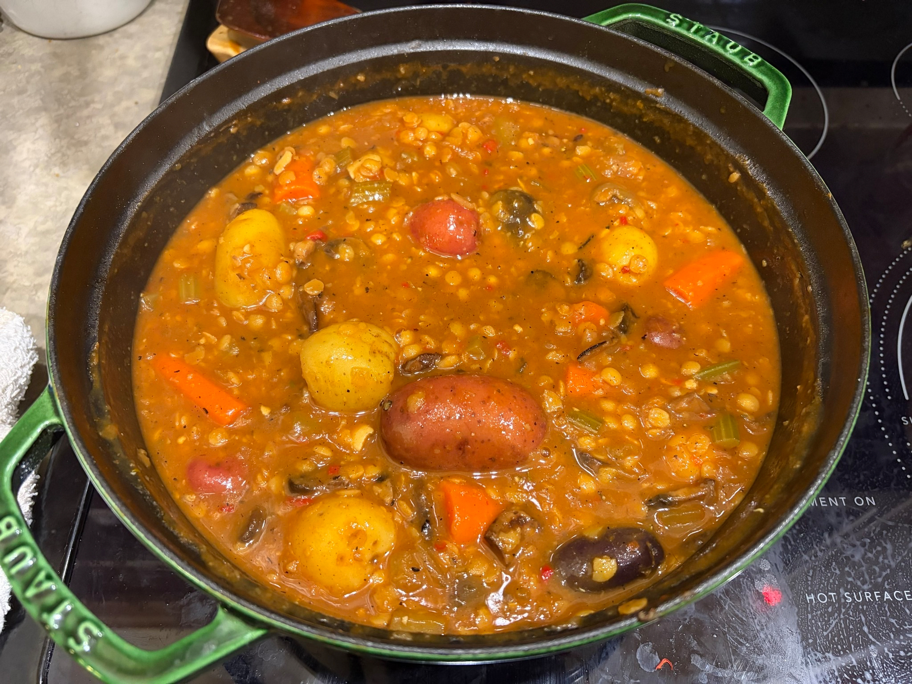 A pot filled with a hearty lentil stew containing potatoes, carrots, tomatoes, and mushrooms is cooking on a stovetop.