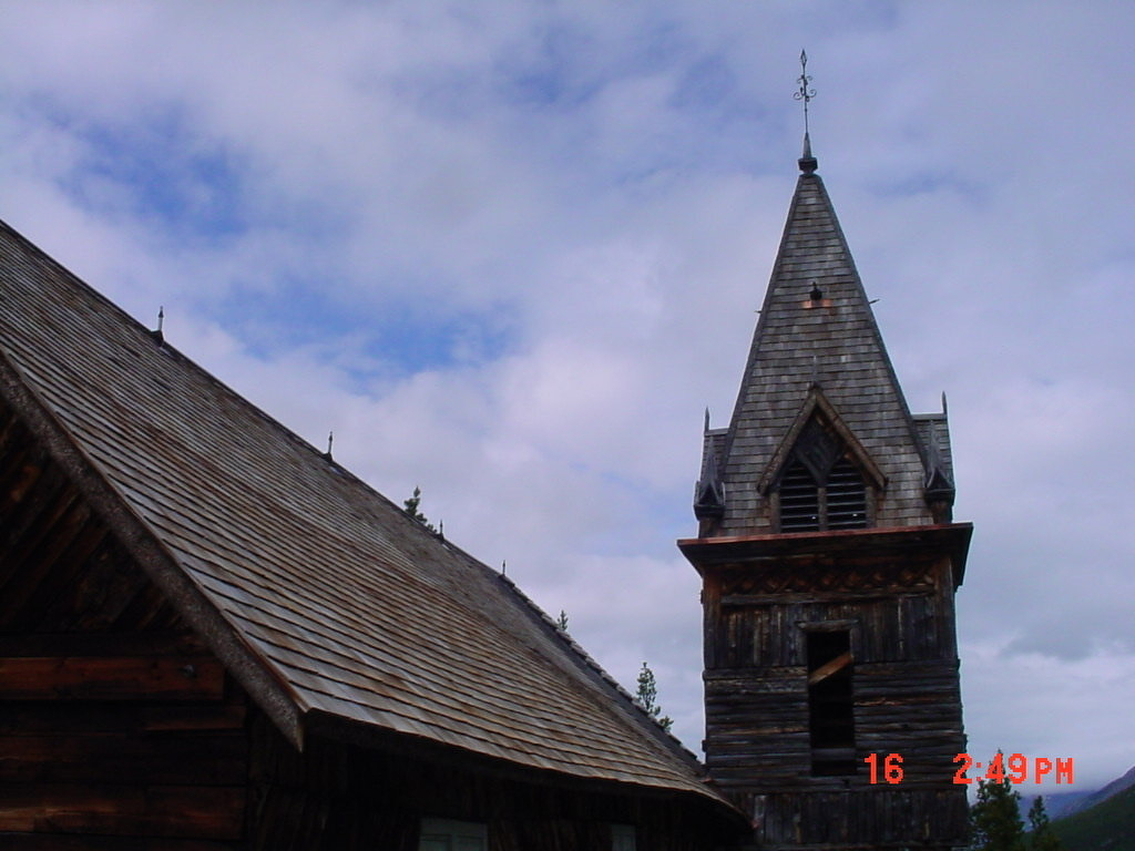 Auto-generated description: A weathered wooden church tower with a steep, shingled roof stands against a partly cloudy sky.